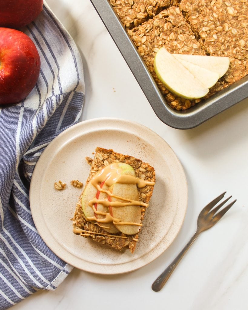 A slice of baked oatmeal served on a side plate topped with apple slices and peanut butter. There is a small fork beside it