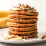 A stack of pancakes topped with banana slices and chopped pecans with a drizzle of maple syrup dripping down it.