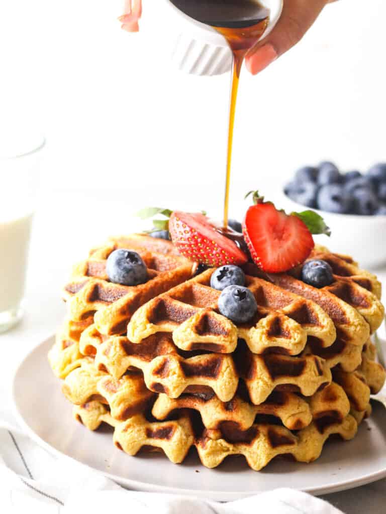 Pouring maple syrup over a stack of vegan protein waffles that are topped with fresh berries.