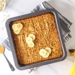 Oatmeal bars in a black square baking pan topped with peanut butter and sliced bananas