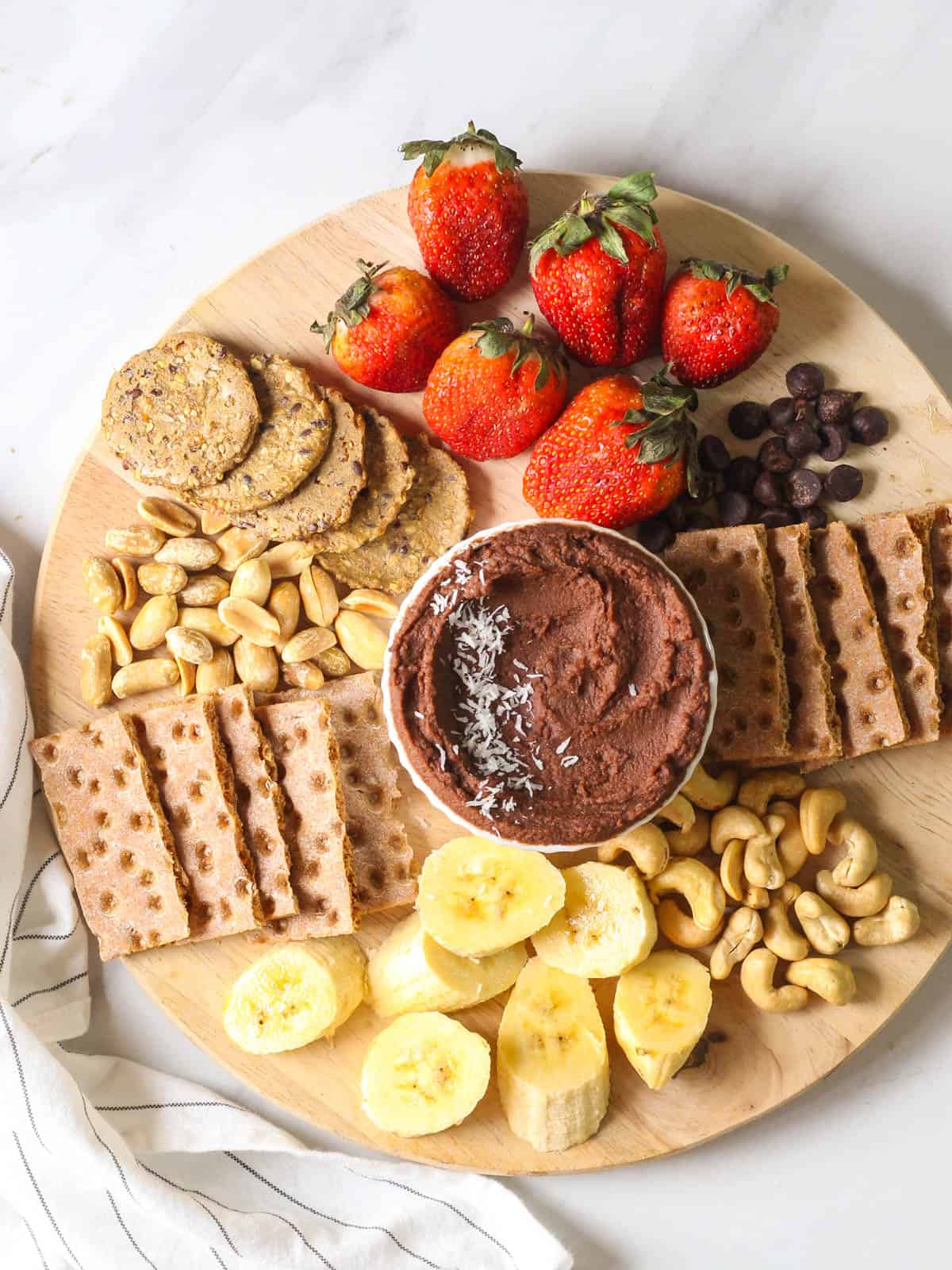 An overhead shot of vegan chocolate hummus served with fruits, nuts and crackers on a wooden chopping board.