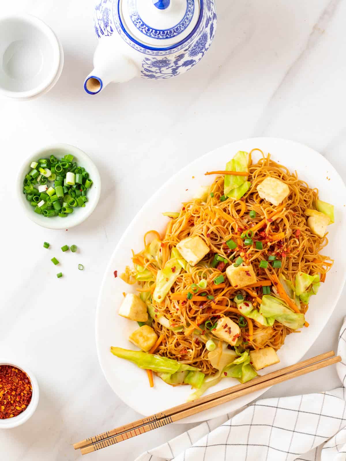 A overhead shot of fried rice vermicelli served in a white plate. There is a plate of green onion and a set of teapot in the background.