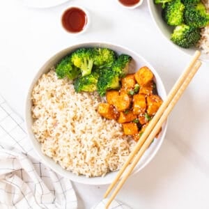 Spicy gochujang tofu served with brown rice and broccoli in a deep plate with a pair of chopsticks on the side.