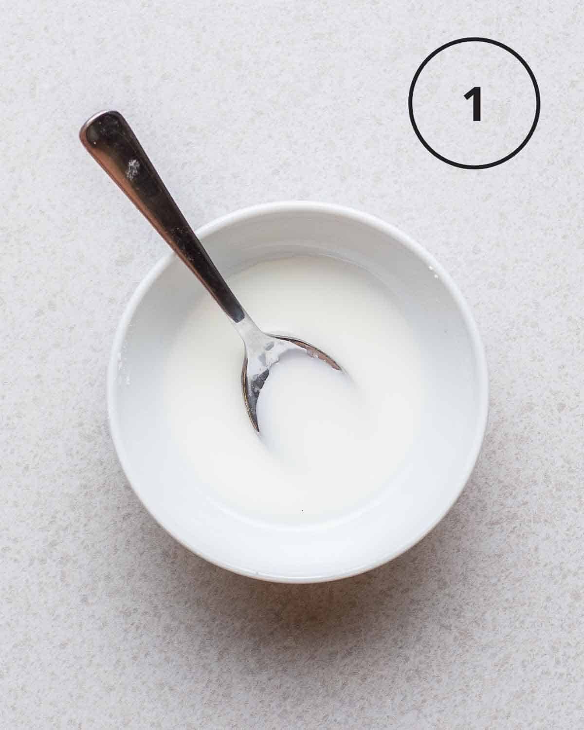 cornstarch slurry in a white bowl with a spoon on a grey background.