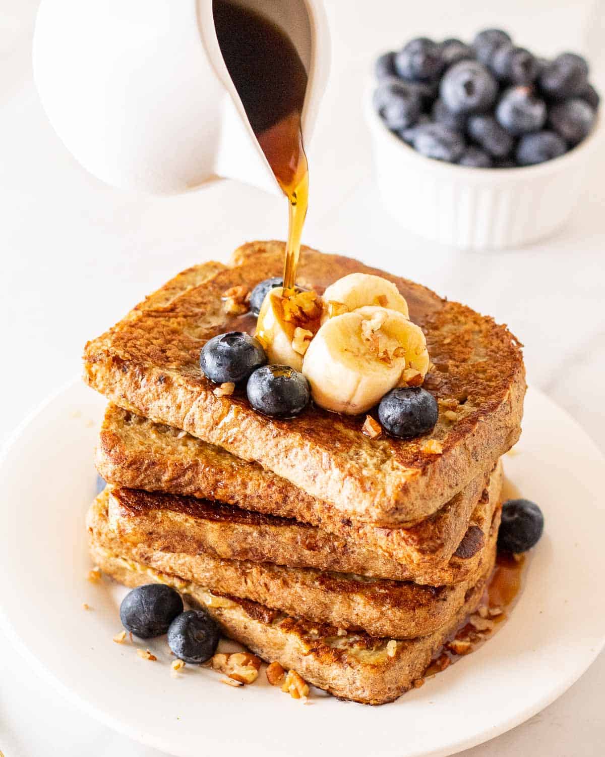 Pouring maple syrup over a stack of eggless French toast. There is a bowl of blueberries in the background.