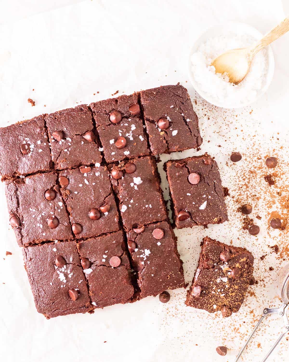 A tray of brownie slices arranged on parchment paper with a small dish of salt flakes besides.