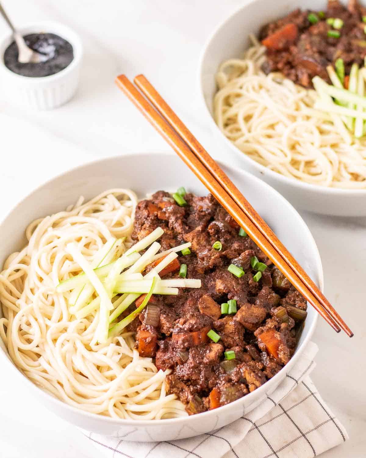 A bowl of vegan jajangmyeon with chopsticks. There is another bowl of noodles and black bean sauce behind.