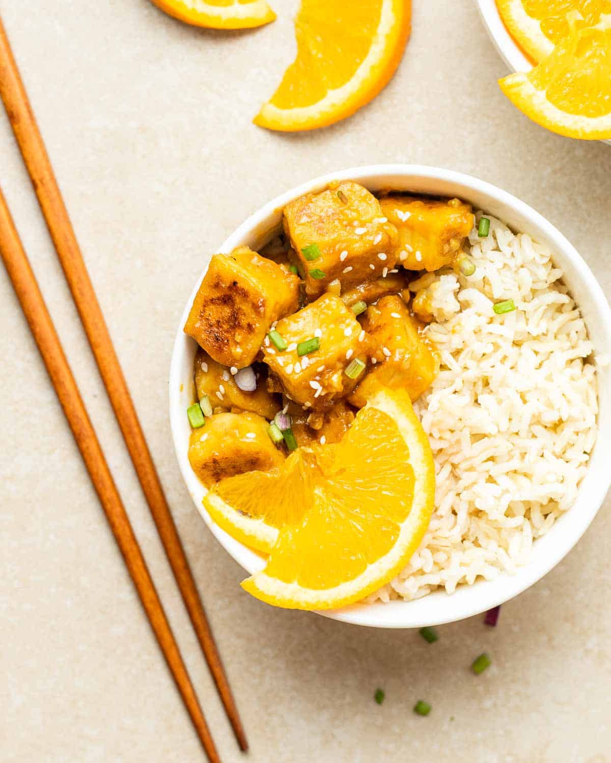 Orange tofu served on a bed of rice topped with scallions and orange slices with chopsticks beside it.