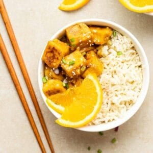Orange tofu served with brown rice and orange slices in a white bowl with a pair of chopsticks beside.