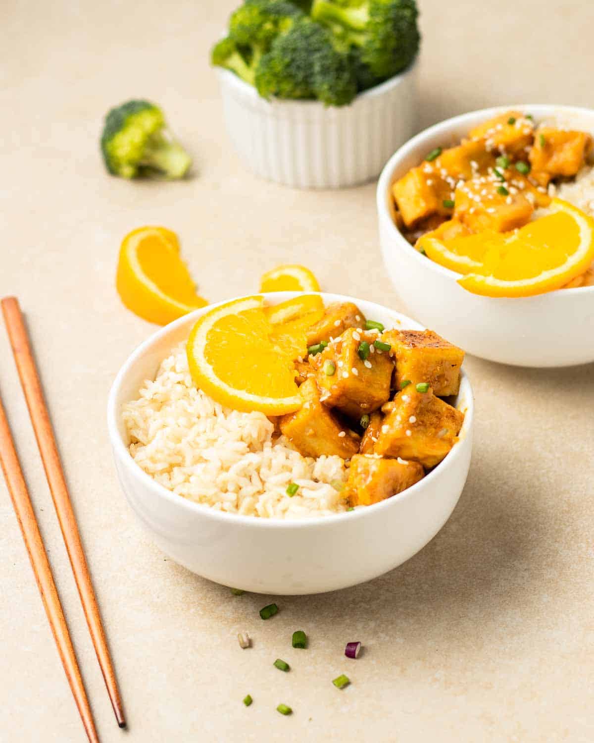 2 bowls of rice served with orange tofu. There is chopsticks and a bowl of broccoli florets in the background.