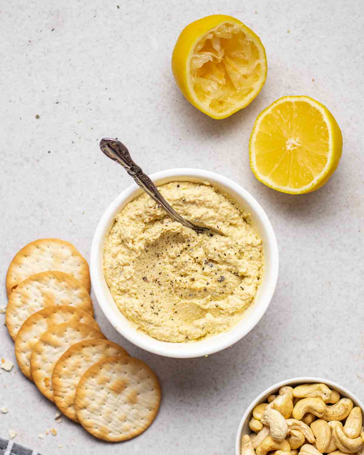 A bowl of dairy-free ricotta with a spoon. There lemon halves, crackers and bowl filled with cashews beside.