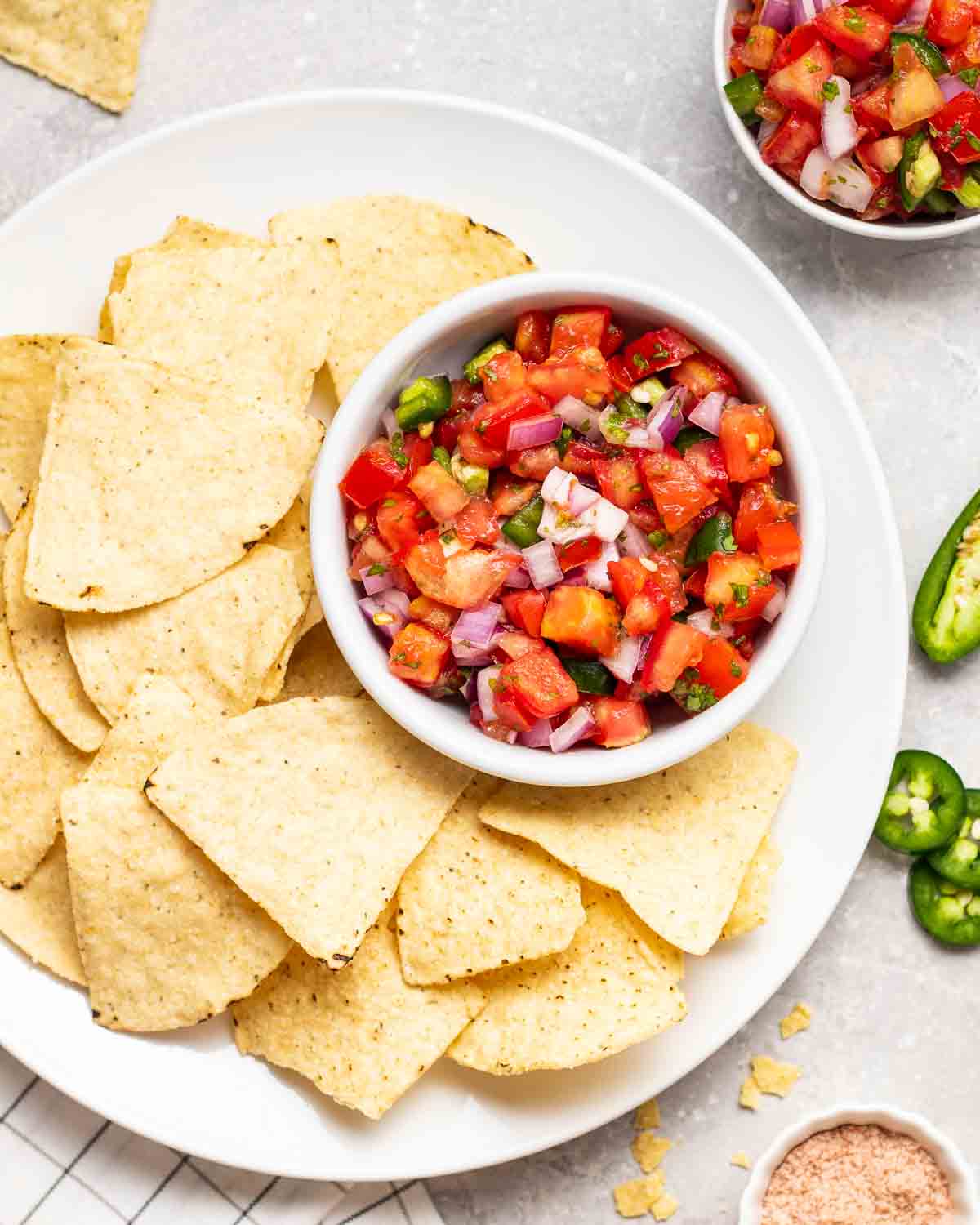 Salsa served with a plate of chips. There are jalapeno peppers and salt in the background. 