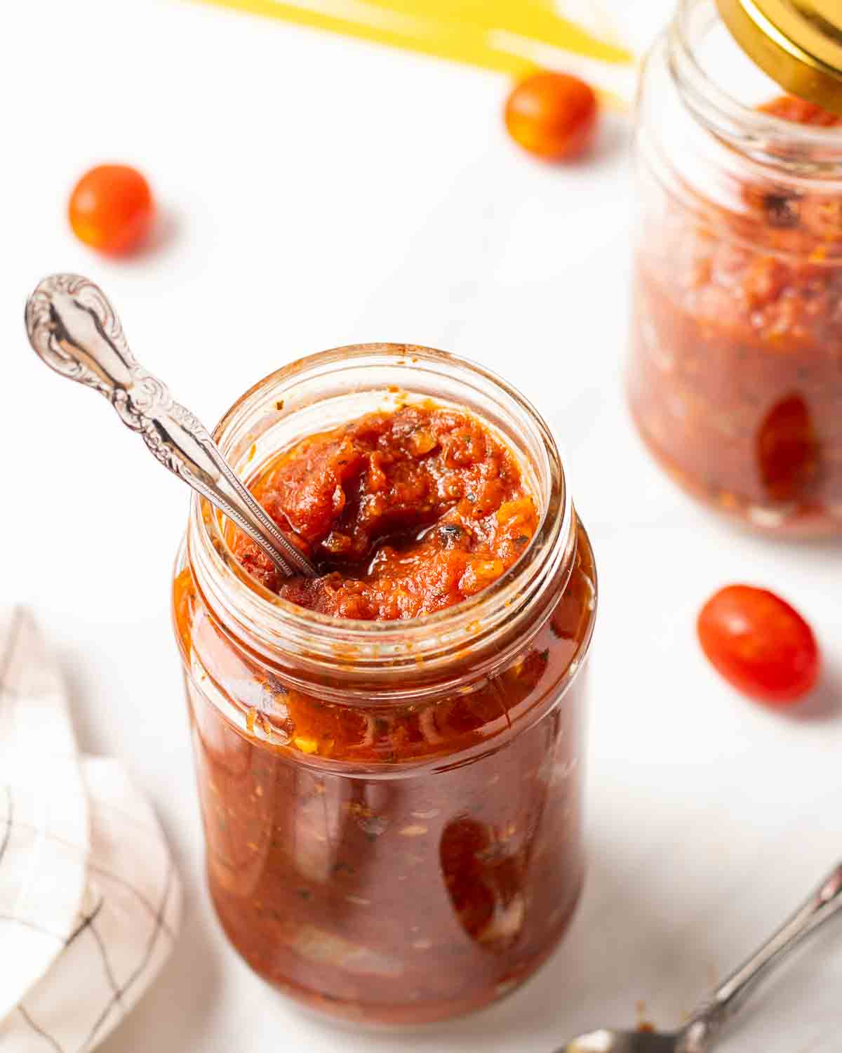 A jar of marinara sauce with spoon. There is another jar of sauce and tomatoes in the background.