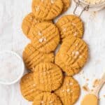 PB2 cookies on a white surface with bowl of sea salt, a spoonful of peanut butter and jar of PB2 beside.