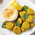 Falafels in a white plate topped with parsley leaves and served with hummus and lemon slices.