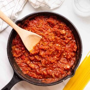 Marinara sauce in skillet with stirrer. There is some spaghetti and glass of water beside.