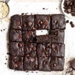 Tray of brownies on a parchment paper with dish of chocolate chips and cup of oats beside.