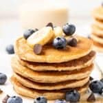 Stack of vegan protein pancakes topped with banana slices, blueberries and chocolate chips with a glass of milk behind it.