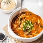 Soup in a white pot with a ladle. There is a bowl of rice behind.