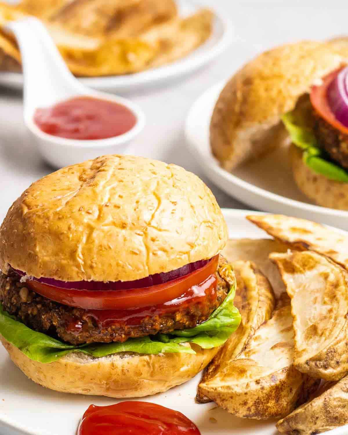Burger served with a side of potato wedges in a white plate with dish of tomato sauce in the background.