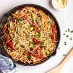 Teriyaki soba noodles in a pan with a dish of sesame seeds, scallions and a wooden spatula beside.