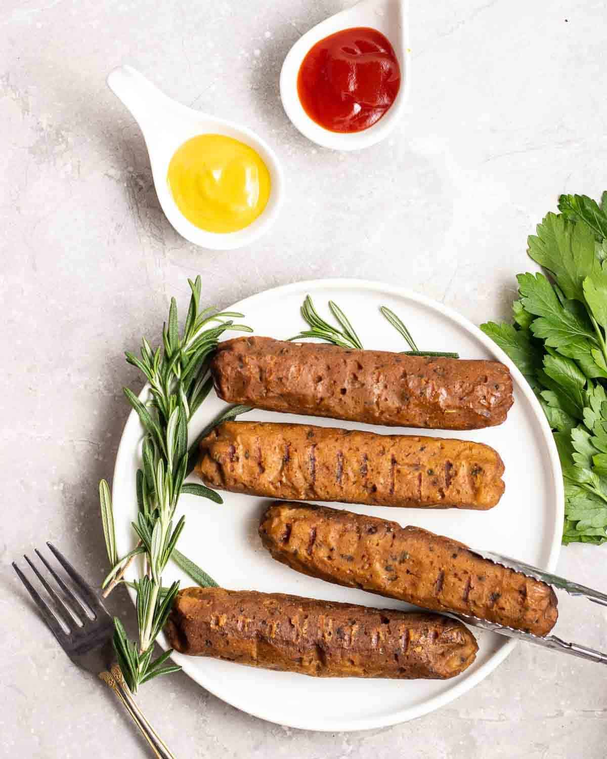 Tongs taking out a piece of sausage from a plate with dish of mustard and ketchup beside.
