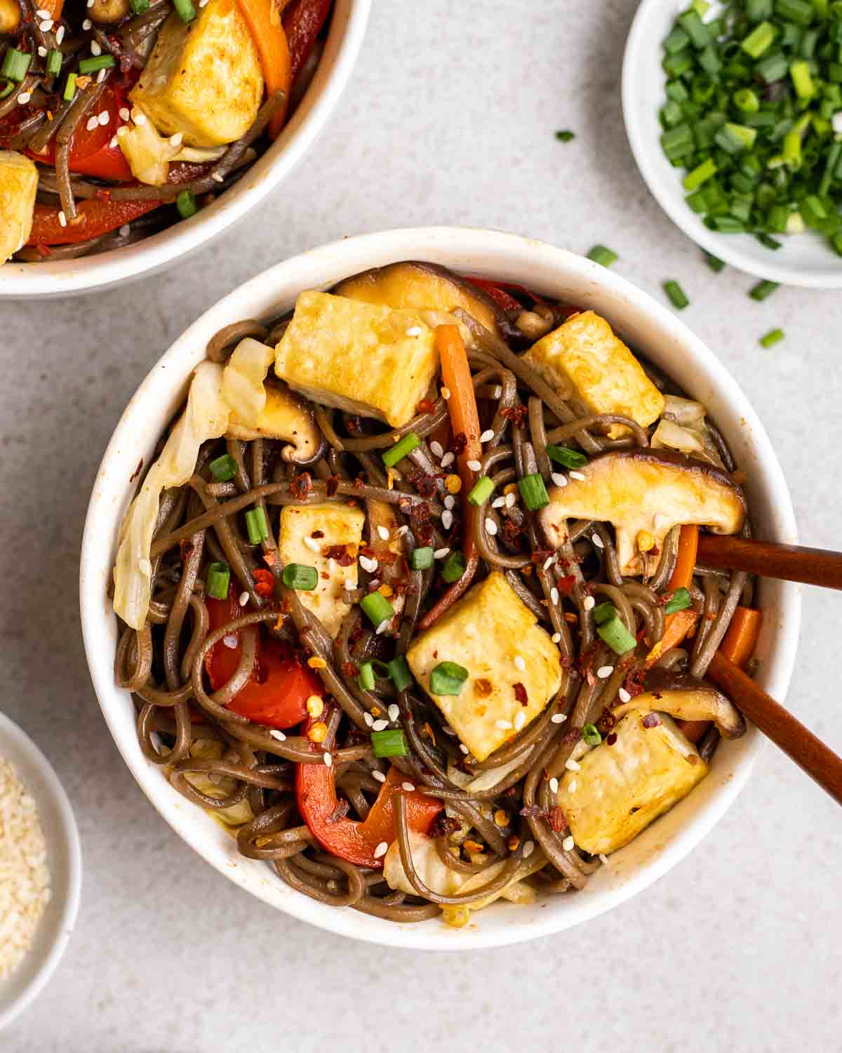 Yakisoba in a white bowl topped with chopped scallions and sesame seeds with chopsticks.
