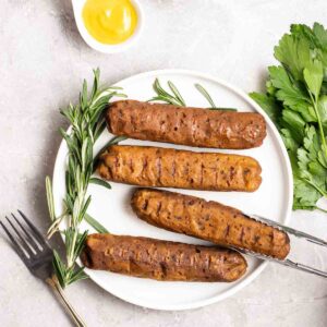Tongs taking out a piece of sausage from a plate with a fork and fresh herbs beside.