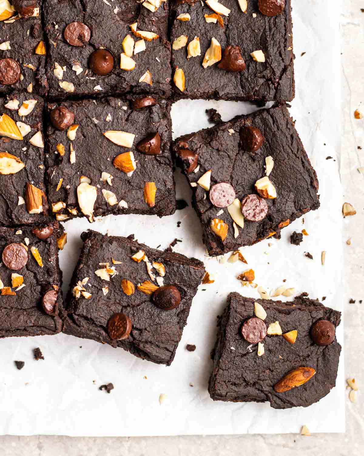 A tray of brownies on a parchment paper topped with chopped almonds and chocolate chips.