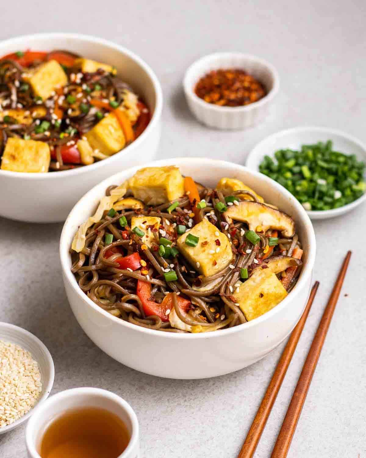 2 bowls of yakisoba with tea, chopsticks, chopped scallions and chili flakes beside.