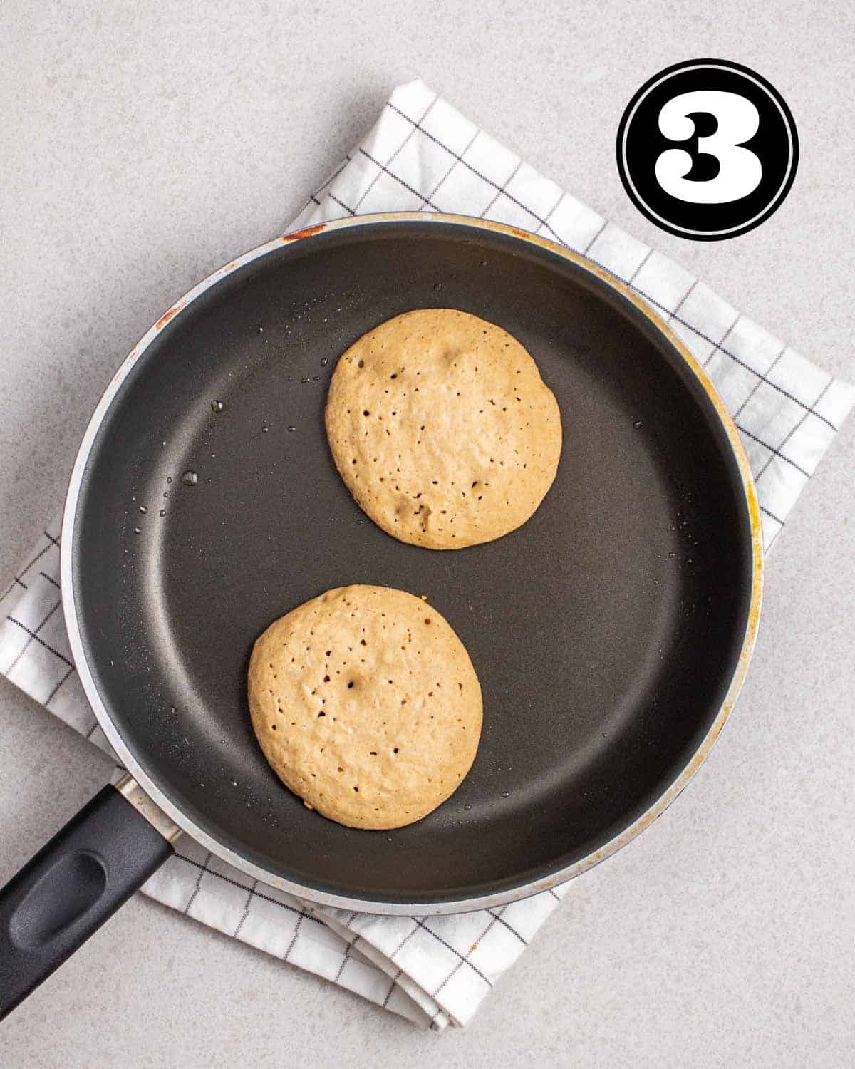 Partially cooked pancakes in a non-stick pan on a white checkered folded teacloth.