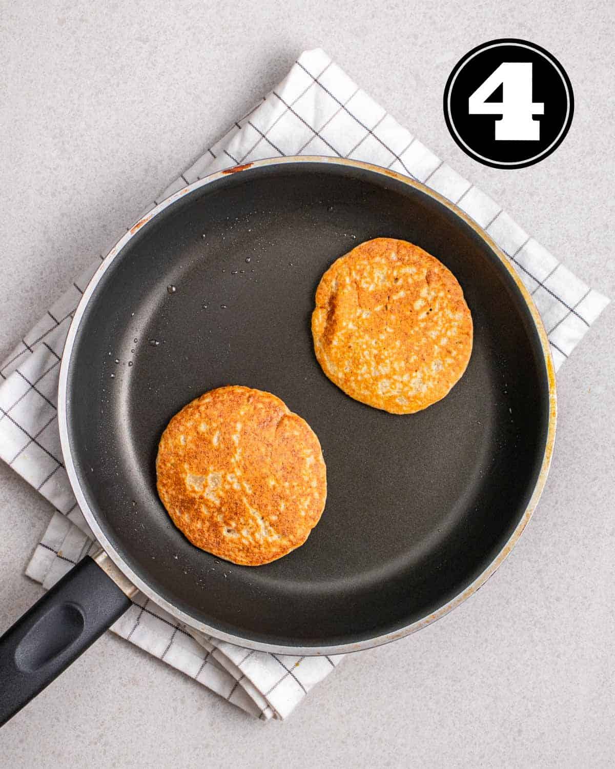 2 cooked pancakes in a non-stick pan on a white checkered teacloth.
