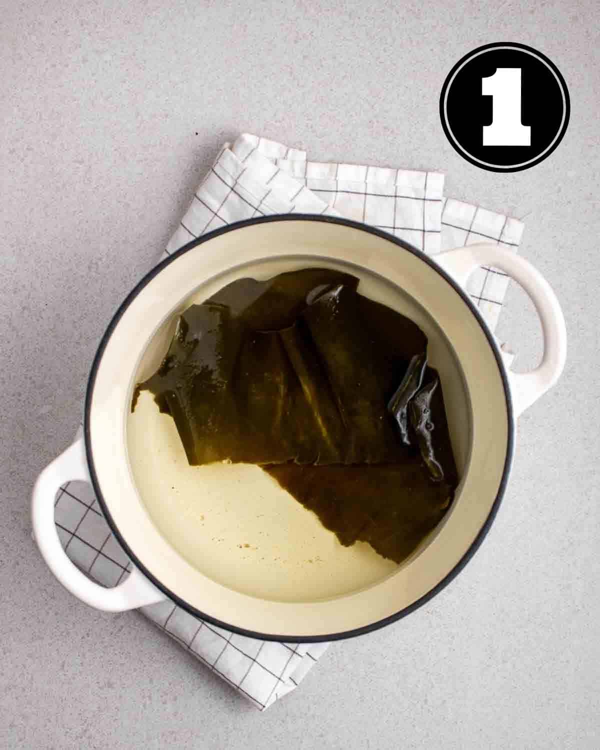 A pot filled with water with kombu inside on a checkered teacloth.