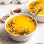 2 bowls of pumpkin potato soup with small dishes of chili flakes and black pepper in the background.