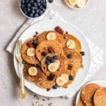 Banana pancakes arranged on a plate with a fork beside topped with blueberries, banana slices and pecans.