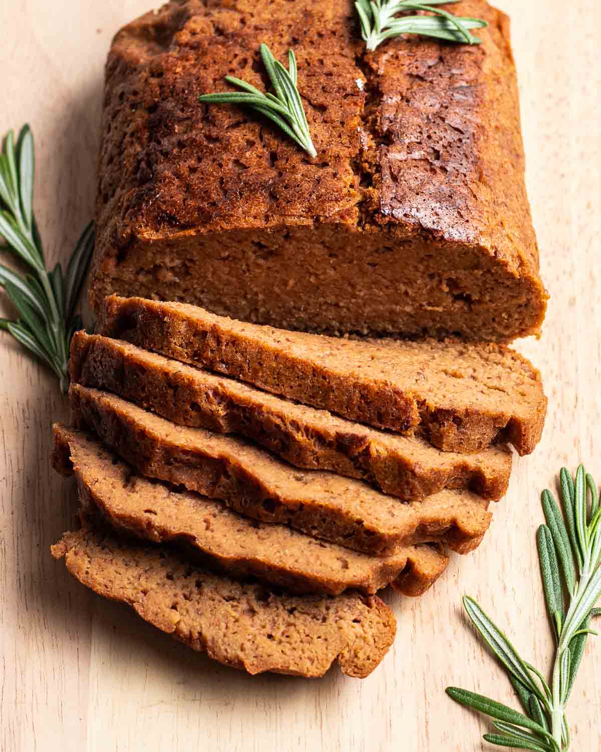 A partially slice seitan roast beef on a wooden board with stems of rosemary beside.