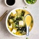A bowl of wakame miso soup served with spoon with chopsticks, chopped green onion and soy sauce beside.