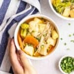 Holding a bowl of tofu vegetable soup with teacloth and chopped green onion beside.