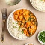 Plate containing sweet potato chickpea curry with rice with a fork, glass of water and dish of chopped cilantro beside.