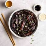 Bowl filled with tteokbokki with chopsticks, cup of tea, soy sauce and sesame seeds beside.