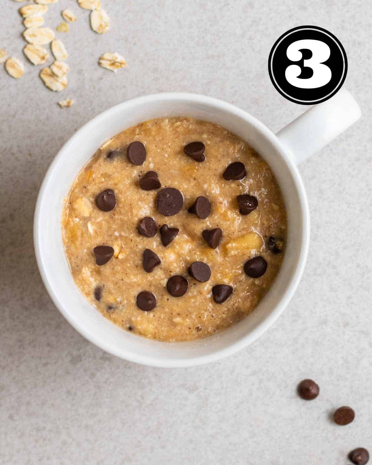 Uncooked mug cake topped with chocolate chips with oats and chocolate chips in the background.