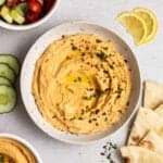 A plate of hummus with cucumber slices, salad, sliced pita and lemon slices beside.