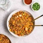 Plate of fried rice with chopsticks beside with glass of water, sambal and chopped green onion in the background.