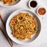 A plate of bami goreng with chopsticks, soy sauce and chili flakes beside.