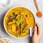 A bowl of vegetable curry served with ceramic spoon with spoonful of curry powder, rice and water beside.