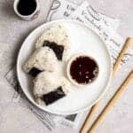 3 onigiri on a plate served with a plate of soy sauce with chopsticks and tea beside.