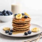 A stack of pancakes on a plate topped with banana and blueberries with forks beside.