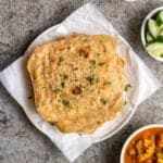A stack of plain paratha on a lined plate with chopped cucumber and curry beside.