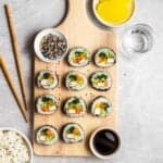 Kimbap arranged on a wooden chopping board with sesame seeds and soy sauce, and chopsticks beside.