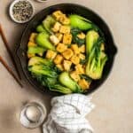 A pan of bok choy tofu with chopsticks, water, and sesame seeds beside.